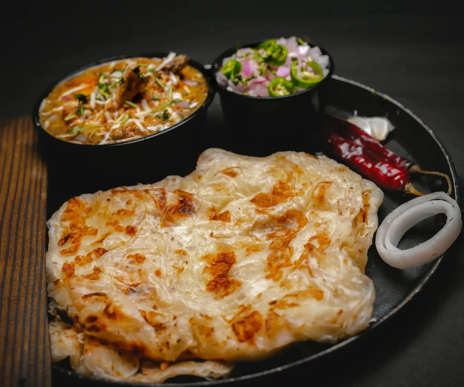 A flaky lachha paratha on a black plate, with a bowl of curry and an onion and chili salad