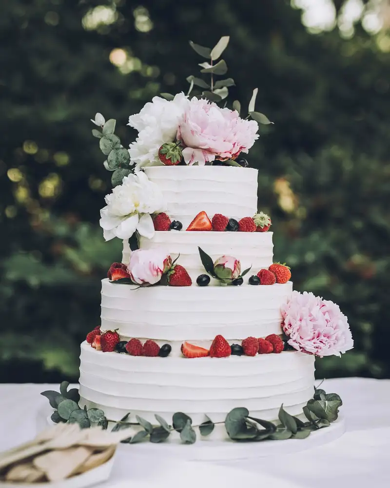 A three-tier white cake with fresh berries, peonies and greenery on a garden table
