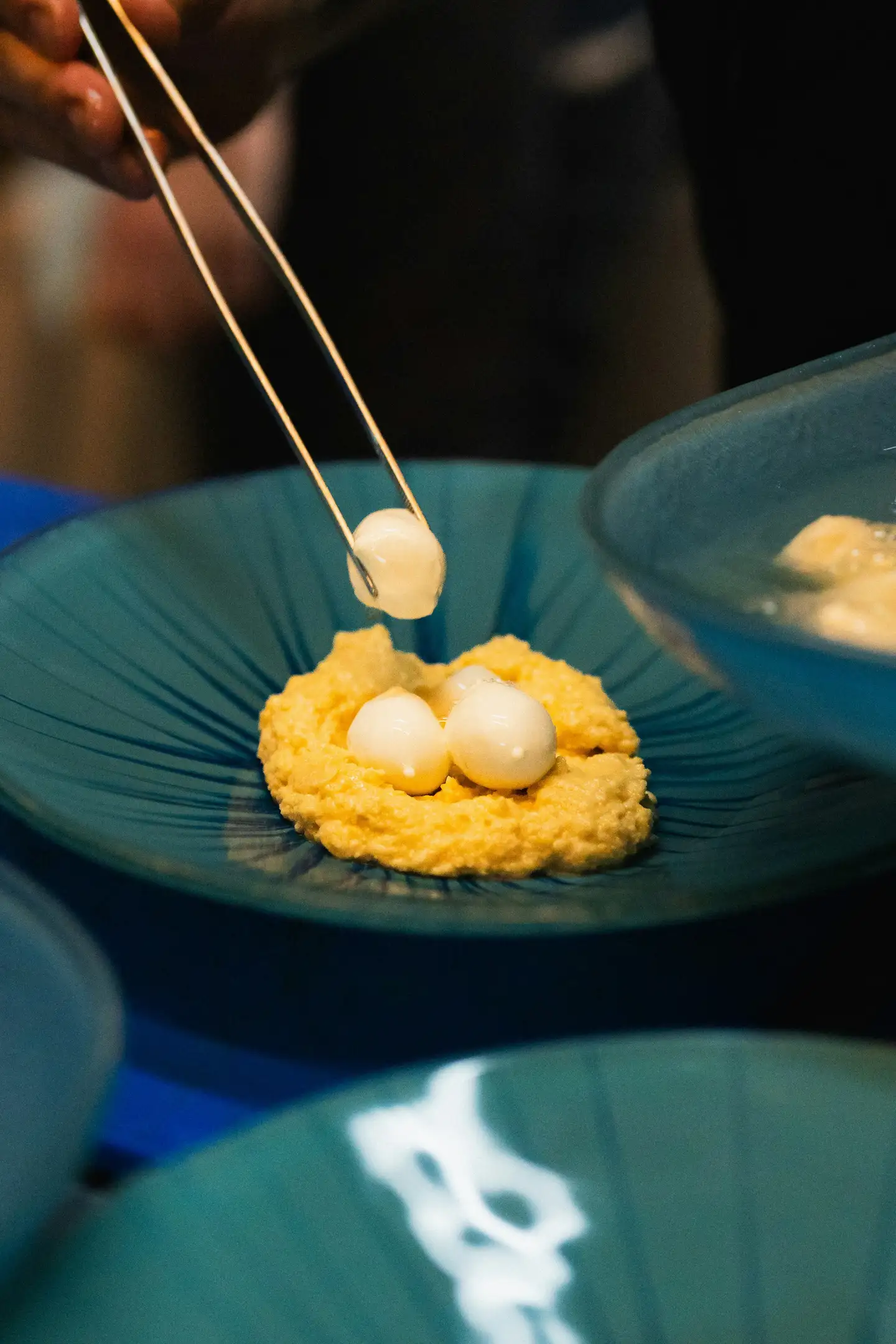 Tweezers setting pearls of cultured cream on a squash purée, on teal plates at the pass