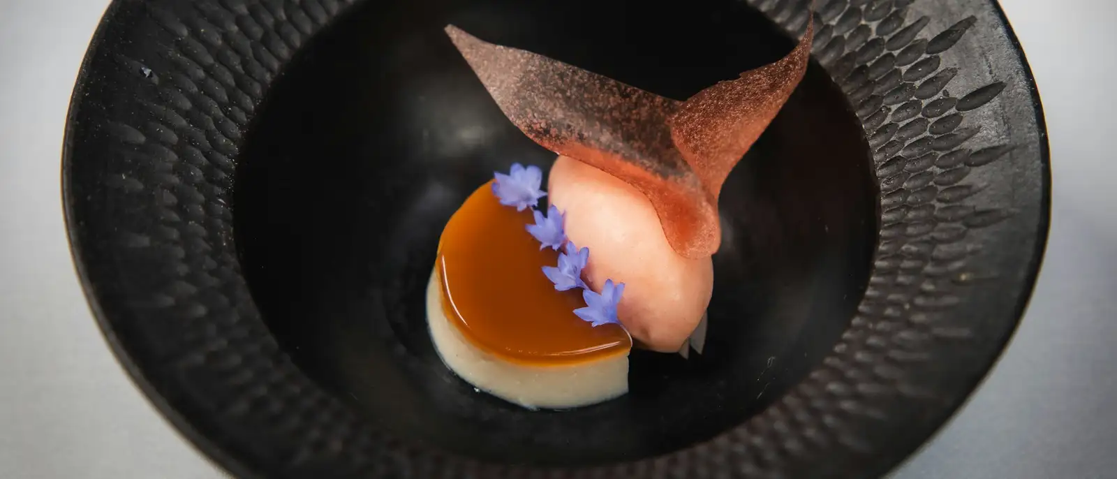 Custard, a pink haskap sorbet and an oat crisp with borage flowers, in a dark ceramic bowl