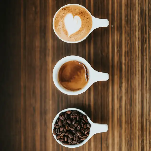Three cups in a row on dark wood: a latte, a cappuccino and a cup of whole beans