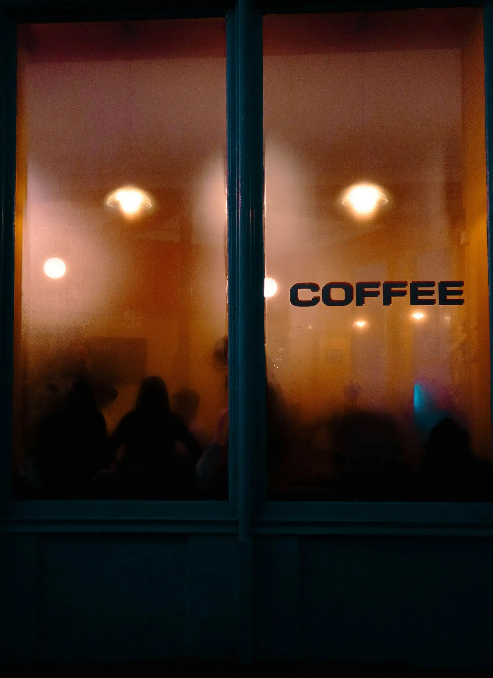 The café window at dusk, warm bulbs glowing inside behind a sign that reads Coffee