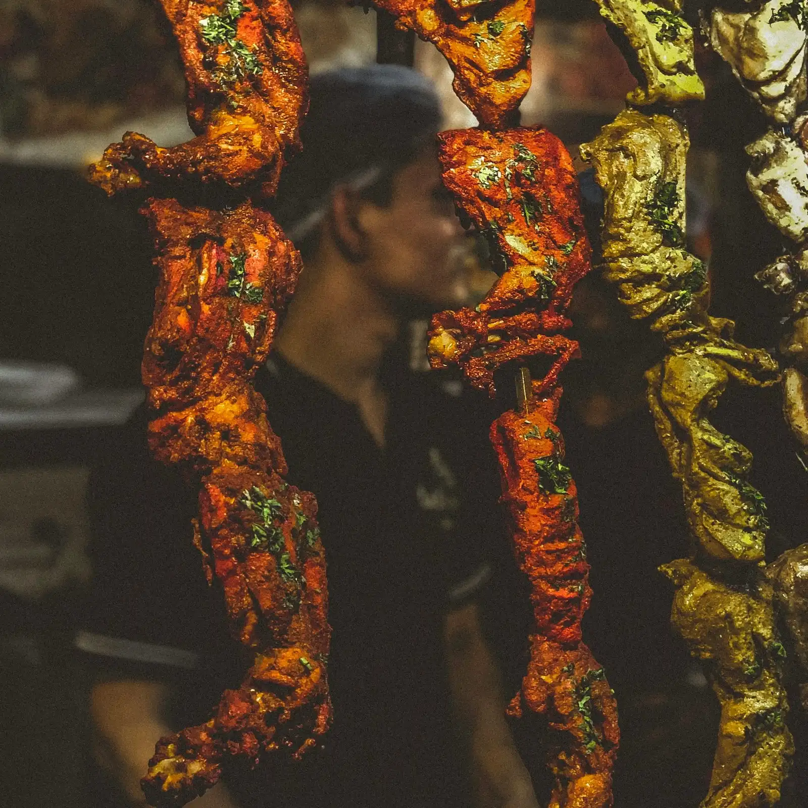 Tandoori chicken glowing red on long skewers, a cook in the warm dark behind them