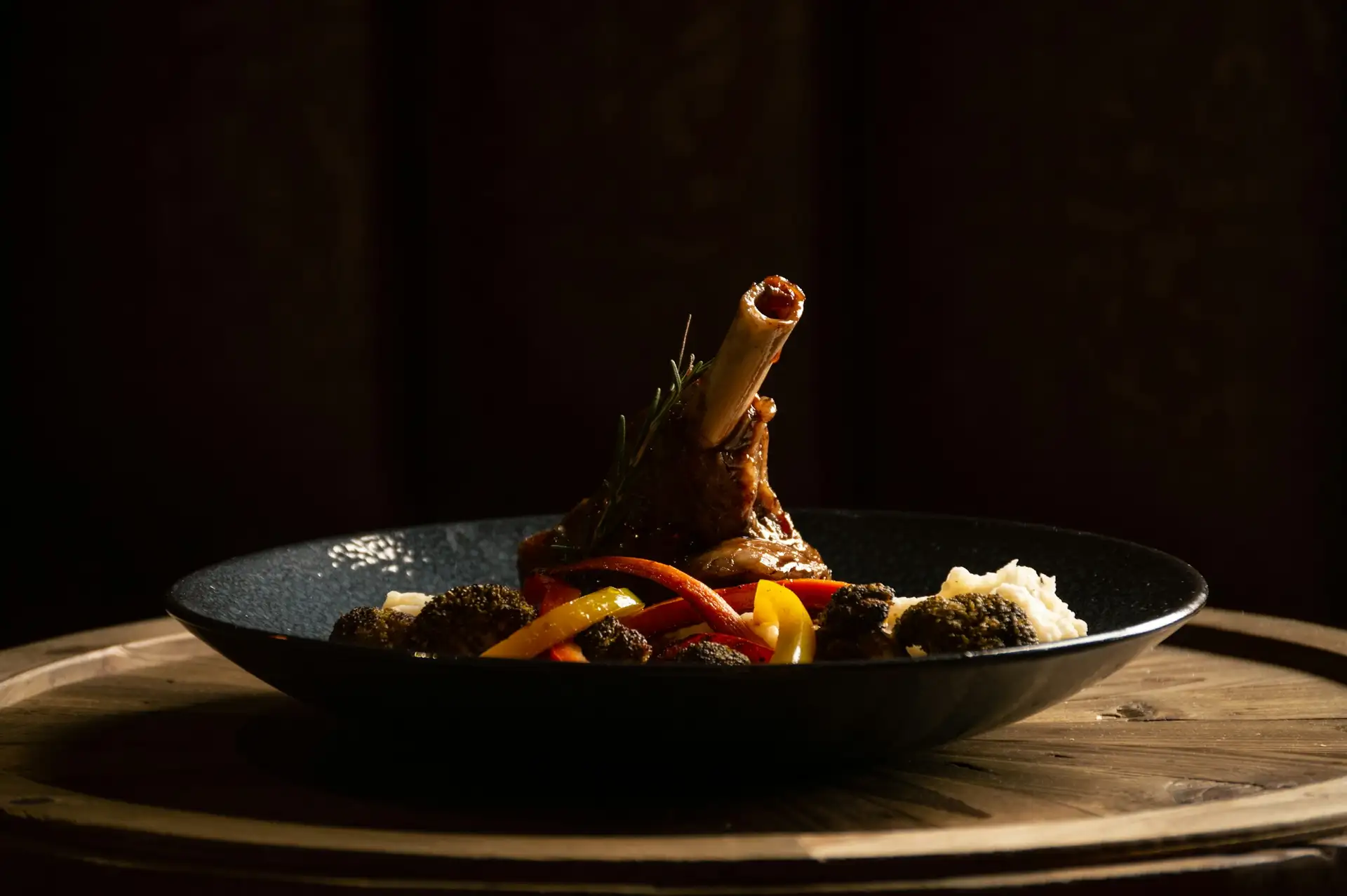 A braised bison shank with charred peppers in a dark bowl, lit low on a wooden board