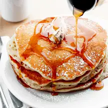 A tall stack of buttermilk pancakes, syrup pouring over a pat of butter, a mug of coffee behind