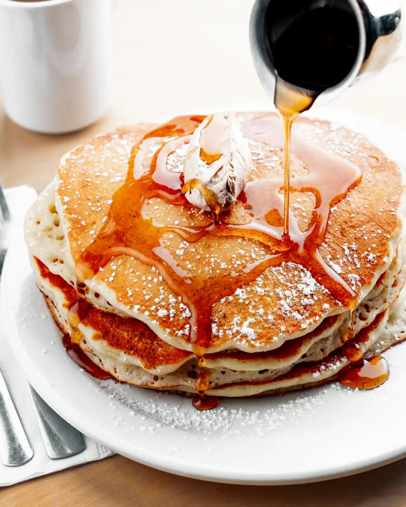 A tall stack of buttermilk pancakes, syrup pouring over a pat of butter, a mug of coffee behind
