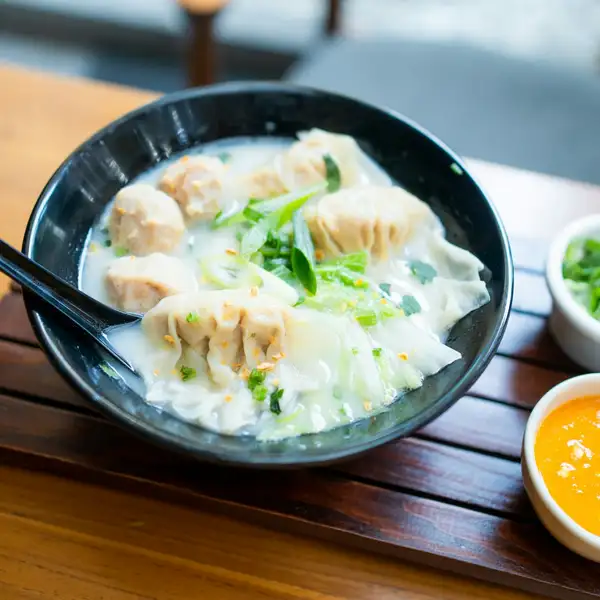 A bowl of wontons in pale broth with greens, small dishes of chopped green onion and chili sauce alongside