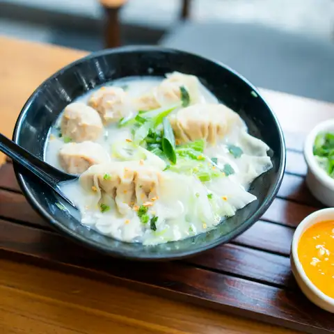 A bowl of wontons in pale broth with greens, small dishes of chopped green onion and chili sauce alongside