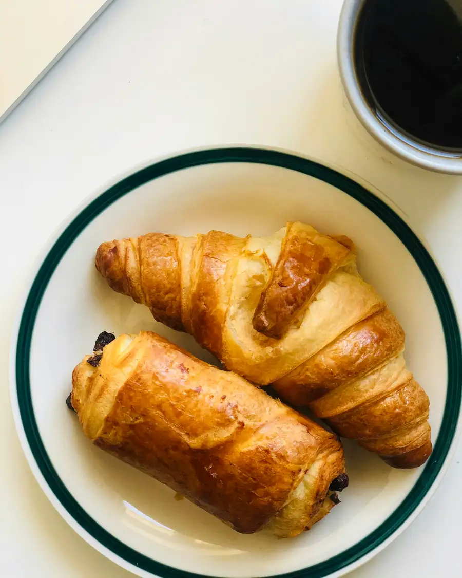 A pain au chocolat and a croissant on a white plate with a green rim, coffee beside