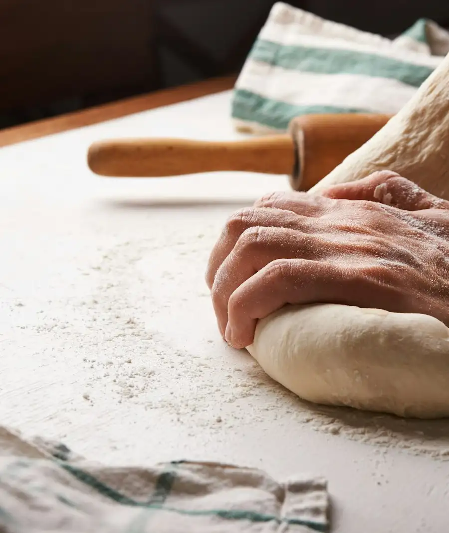 Hands kneading a round of dough on a floured counter, a rolling pin behind