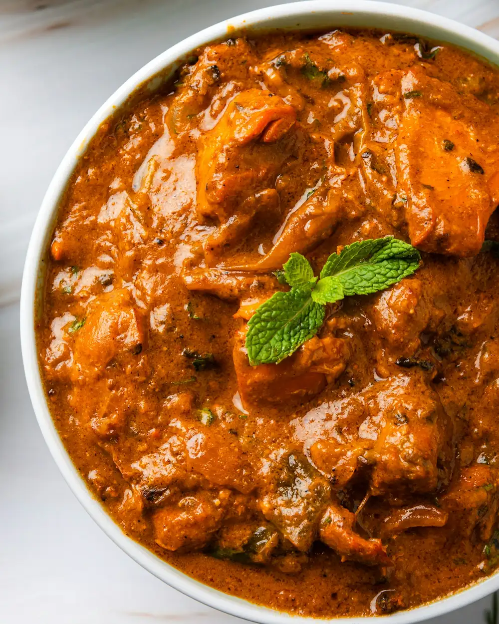 Butter chicken in a white bowl, glossy orange sauce topped with a sprig of mint