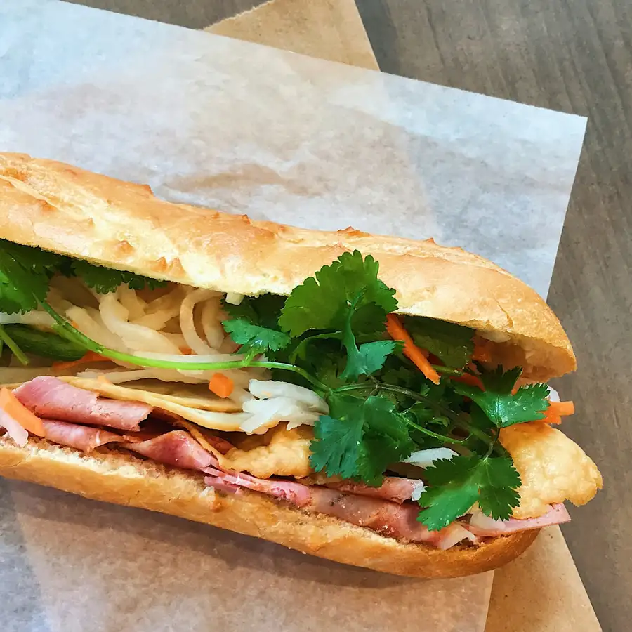 A bánh mì split open with ham, pickled carrot, cucumber and cilantro, on white paper