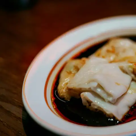 Silky rice noodle rolls in a pool of sweet soy sauce on a white plate