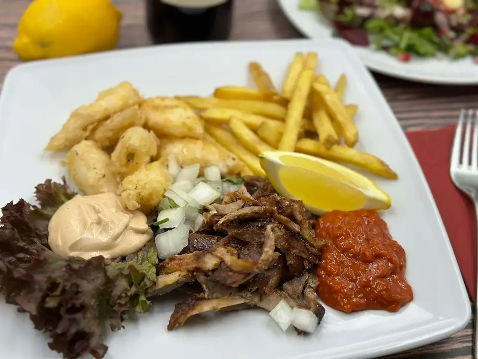Shaved shawarma on a white plate with fries, crispy fried bites, garlic sauce, red pepper dip and a lemon wedge