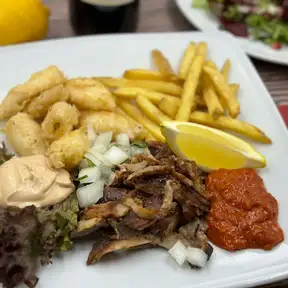 Shaved shawarma on a white plate with fries, crispy fried bites, garlic sauce, red pepper dip and a lemon wedge