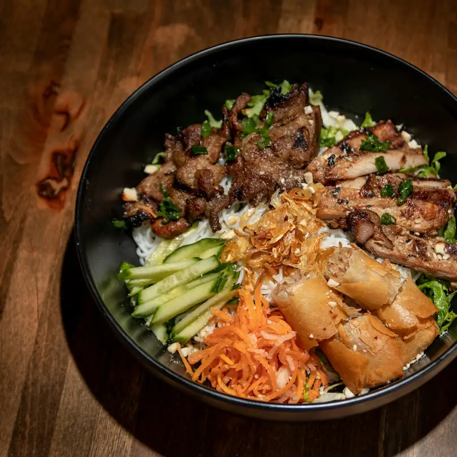 Vermicelli topped with grilled pork, cut spring rolls, cucumber and pickled carrot in a black bowl