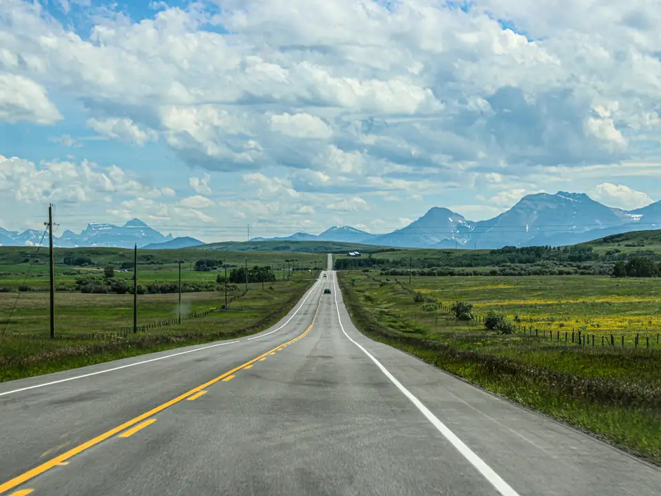 A two-lane highway running straight across green prairie toward the mountains, under a big sky