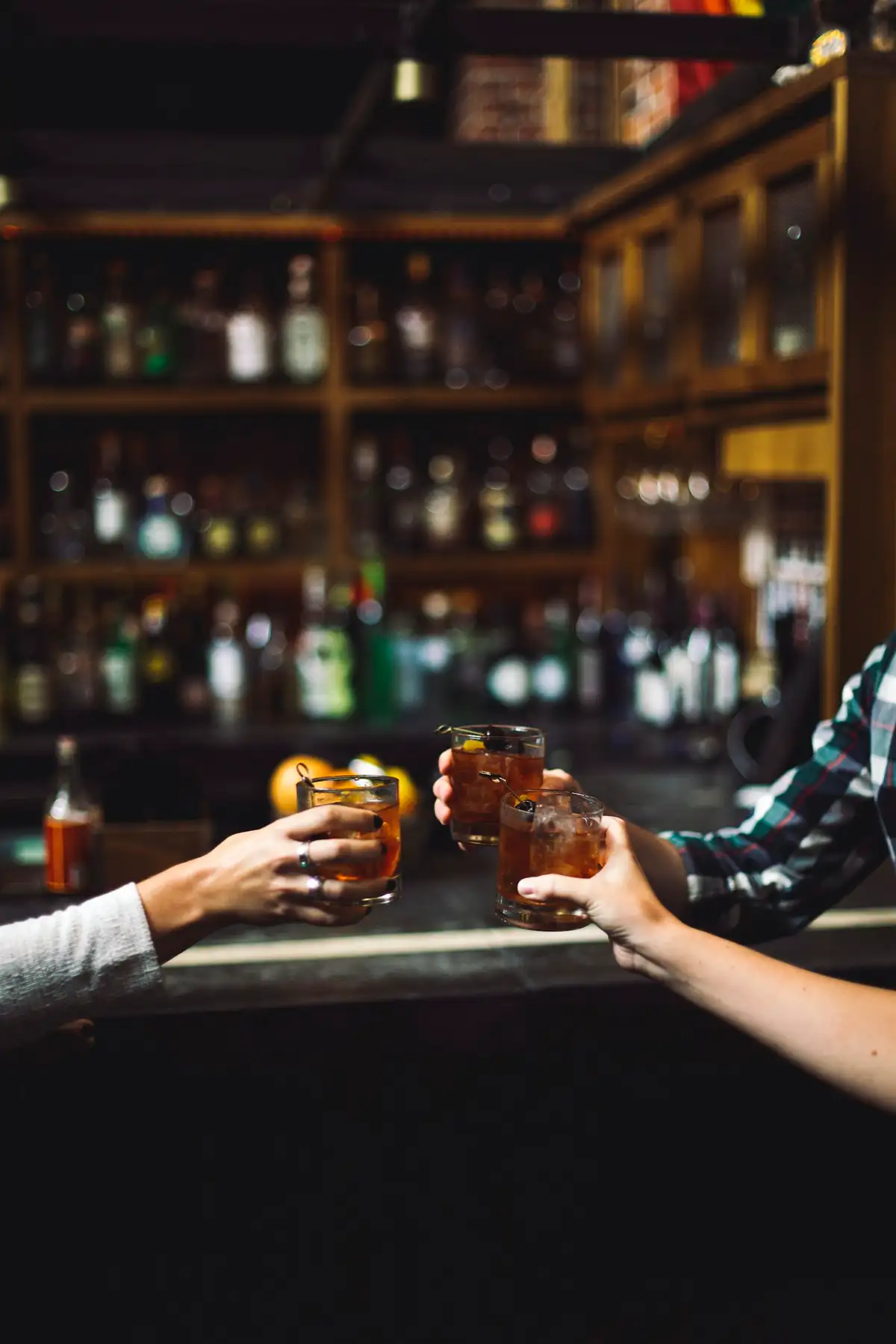 Friends clinking Old Fashioneds across the bar, shelves of bottles glowing behind